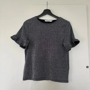 Glittrig topp från NA-KD  - Snygg glittrig topp från nakd i en mörkgrå nyans. Den har korta ärmar med volangdetaljer som ger en extra touch. Perfekt för festliga tillfällen eller när du vill glittra lite extra i vardagen. Passar bra till både jeans och kjol!