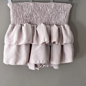 Beige volangkjol - Säljer en superfin beige kortkjol med volanger. Den har en smockad midja som ger en bekväm och flexibel passform. Perfekt för sommardagar eller en avslappnad fest. Kjolen är i nyskick och redo för nya äventyr! Har inbyggda shorts, passar xs-s. Passar så bra till en stickad tröja!!