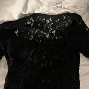 Svart spetstopp  - Säljer en elegant svart spettopp med vackra detaljer. Toppen har en knytning framtill och är perfekt för festliga tillfällen. Den är i nyskick och har en romantisk känsla med sitt fina spetsmönster.