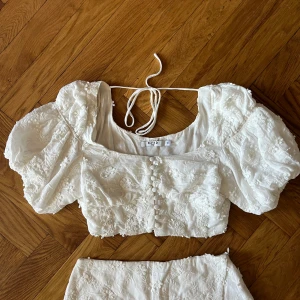 NA-KD X Chloe Schuterman vit topp - NA-KD X Chloe Schuterman vit topp  Blomster broderier och knappar framtill. Croppad med puff armar  Säljer även tillhörande kjol, går att sammköpa.  Toppen skick,använd vid få tillfällen  Storlek 38 men passar en S