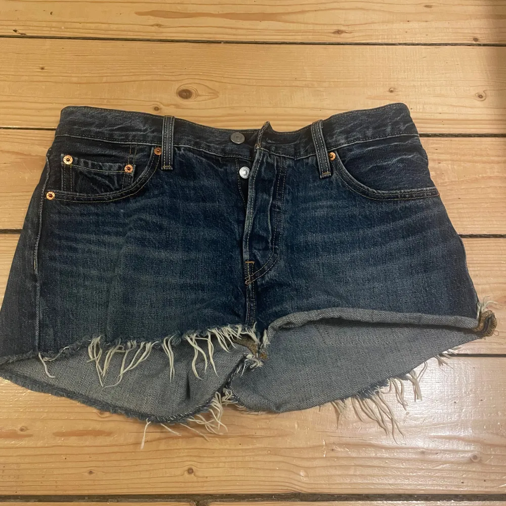 Säljer ett par snygga jeansshorts från Levi's. De är mörkblå med en fransig kant nertill för en cool och avslappnad look. Perfekta för sommaren och festivaler! De har en klassisk femficksdesign och knappar framtill.. Shorts.