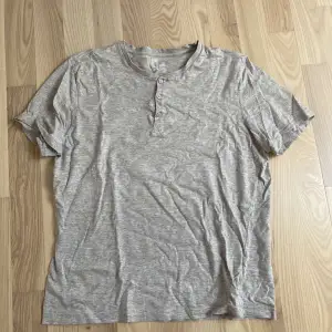 Säljer en grå t-shirt från H&M i storlek M. Den är kortärmad och har tre knappar vid halsen för en avslappnad look. Perfekt för vardagsbruk och gjord i mjukt bomullsmaterial.