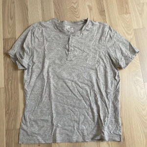 Grå t-shirt från H&M - Säljer en grå t-shirt från H&M i storlek M. Den är kortärmad och har tre knappar vid halsen för en avslappnad look. Perfekt för vardagsbruk och gjord i mjukt bomullsmaterial.