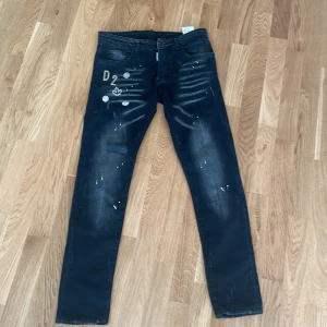 Svarta jeans från Dsquared2 - Säljer ett par svarta slim jeans från Dsquared2. De har en cool, sliten look med vita färgstänk och broderade detaljer på framsidan. Baktill finns en stor etikett med texten 'Caten'. Jeansen är tillverkade i Italien och har en modern, stilren design. Perfekta för en trendig vardagslook!