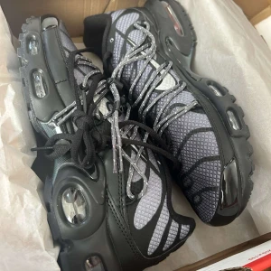 Svarta och grå Nike sneakers - Säljer ett par snygga Nike sneakers i svart och grått. De har en cool design med synliga luftkuddar i sulan och ett mönstrat mesh-tyg på ovansidan. Skorna har snörning och ser riktigt bekväma ut för både vardag och träning.