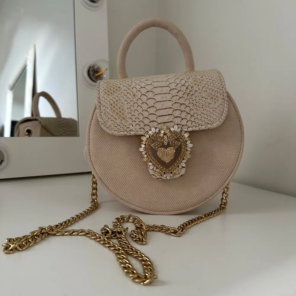 Säljer en superfin beige rund handväska med en lyxig guldkedja. Väskan har ett snyggt ormskinnsmönster på locket och en dekorativ hjärtformad detalj framtill. Perfekt för att ge din outfit en elegant touch! Väskan är köpt utomlands för några år sen🙂. Väskor.