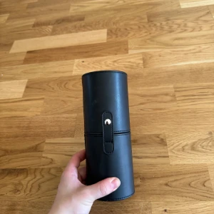 Svart cylinderformad väska - Säljer en stilren svart cylinderformad väska i syntetmaterial. Perfekt för att förvara småsaker eller som en unik accessoar som sminkborstar. Väskan har en enkel design med en knappdetalj för stängning.