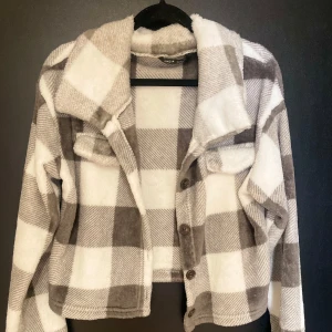 Rutig fleecejacka från Shein - Säljer en supermysig och trendig rutig fleecejacka från Shein. Den är i en snygg geå och vit färgkombination. Jackan har långa ärmar och knappar framtill. Perfekt för höst och vår när du vill vara både varm och stilren. Den är i storlek S och passar perfekt för en casual look.