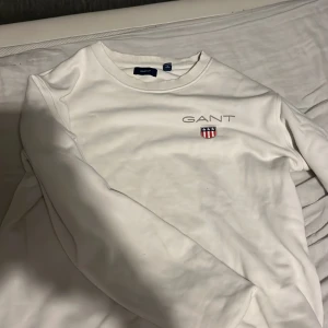 Vit tröja från GANT - Säljer en stilren vit tröja från GANT med broderad logotyp och emblem på bröstet. Tröjan har en rund halsringning och långa ärmar, perfekt för en avslappnad look. 16 år står det att man ska kunna ha den men den är mindre i storleken, 176 cm 