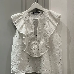Vit blus med volangdetaljer - Säljer en söt vit blus från Zara med volangdetaljer och broderat mönster. Blusen är ärmlös och har en luftig passform, perfekt för varma dagar. Den har en rund halsringning och en liten öppning framtill. Det är storlek medium men passar som S.