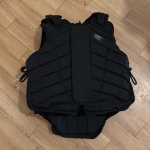 Svart skyddsväst från CRIV - Säljer en svart skyddsväst från CRIV med justerbara sidoband och vadderad design. Västen har en dragkedja framtill och är perfekt för aktiviteter som kräver extra skydd.