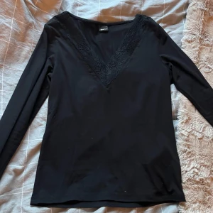 Svart långärmad topp med spetsdetaljer - Snygg svart långärmad topp från gina med spetsdetaljer längs den v-ringade halsen. Perfekt för en stilren look. Knappt använd, inga defekter💗