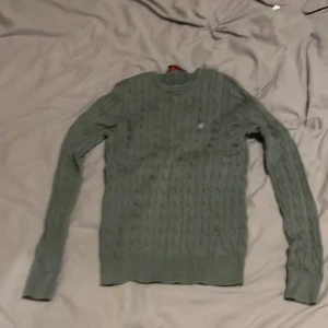 Grön kabelstickad Morris tröja  - Säljer en stilren grön kabelstickad tröja med rund hals och ett diskret broderat emblem på bröstet. Perfekt för kyligare dagar.