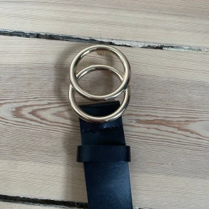 Svart bälte med gulddetaljer från H&M - Snyggt svart skärp från H&M med dubbla guldringar som spänne! Mer av ett högmidjat elr midwaist skärp beroende på hur stor man är