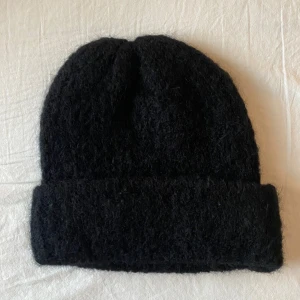Svart stickad mössa från NA-KD i alpacka blend - Snygg och stilren svart stickad mössa från NA-KD. Perfekt för att hålla värmen under kyliga dagar. Den har en klassisk design med uppvikt kant och är tillverkad i en alpacka blend. Helt ny och oanvänd.
