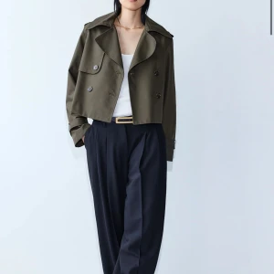 Olivgrön croppad trenchcoat - Snygg olivgrön croppad trenchcoat med dubbelknäppning och bred krage. Perfekt för en stilren look.