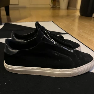 Svarta sneakers från Axel Arigato - Snygga svarta sneakers från Axel Arigato med dragkedja framtill. Skorna har en stilren design med vit sula och är tillverkade i mocka och är i bra skick. Pris kan diskuteras vid snabb affär!!!
