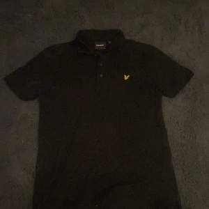 Svart pikétröja från Lyle & Scott - Säljer en stilren svart pikétröja från Lyle & Scott med det klassiska gula logotypen på bröstet. Tröjan har korta ärmar och en bekväm passform, perfekt för en avslappnad look.