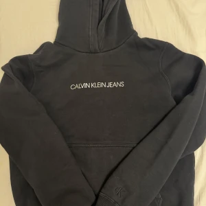 Svart hoodie från Calvin Klein Jeans - Snygg svart hoodie från Calvin Klein Jeans med logga på bröstet. Perfekt för en avslappnad stil. Den har en klassisk huva och långa ärmar. Köpt för 1000kr kan användas till både tjejer och killar, barn tom. 