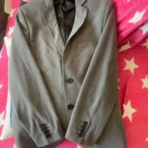 Grå kavaj från Sergio Rossi - Snygg grå kavaj från Sergio Rossi i storlek 48. Tillverkad av polyester, viskos och elastan för en bekväm passform. Kavajen har klassisk design med två knappar framtill och fyra dekorativa knappar vid ärmsluten.