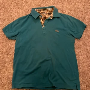 Turkos pikétröja från Burberry - Snygg turkos pikétröja från Burberry med rutigt mönster på insidan av kragen. 9/10 skick, har används varsamt. Kontakta oss om du undrar något!!