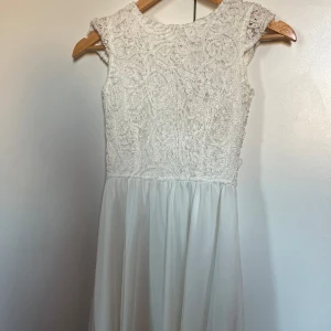 Vit klänning - Elegant vit klänning med spetsdetaljer på överdelen och en flytande kjol. Klänningen har en v-ringad rygg med knappar och är ärmlös, vilket ger en romantisk känsla.