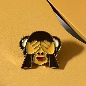 Guld och svart apa-emaljpin - Söt emaljpin i form av en apa som täcker ögonen. Pinnen har en guldig kant och detaljer i svart, beige och röd. Hämtas i Hornstull i Stockholm eller skickas mot fraktkostnad. 🙈