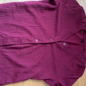 BUDA PRIS: Vinröd blus med knappar - Snygg vinröd blus med en texturerad yta och knappar framtill. Perfekt för att ge en färgklick till din outfit. Blusen har en elegant design med långa ärmar och en bekväm passform.
