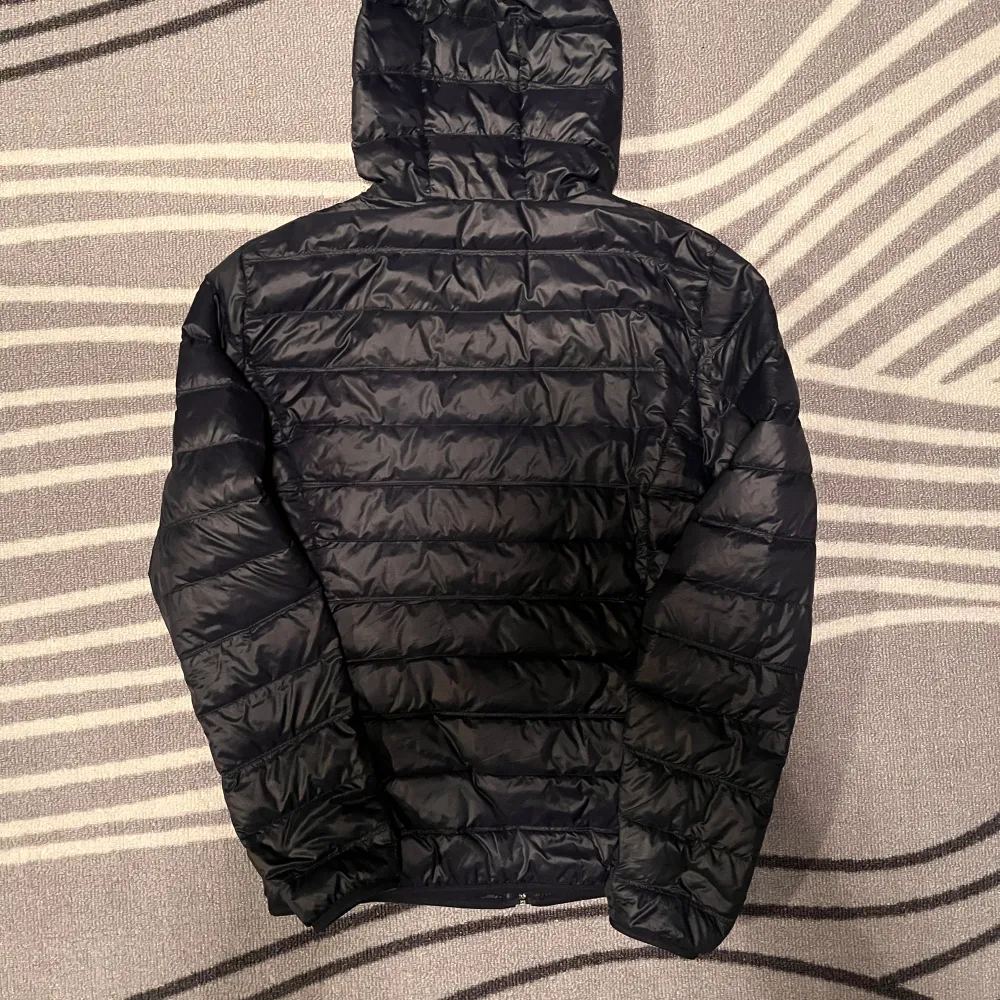 Snygg svart pufferjacka från EA7 Emporio Armani med huva och dragkedja. Perfekt för kyligare dagar med sin quiltade design som håller värmen. Jackan har en stilren look med EA7-logga på bröstet.. Takit.