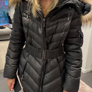 Svart dunjacka från Bark of Sweden - Snygg svart dunjacka från Bark of Sweden med äkta pälskantad huva och dragkedja. Jackan har ett bälte i midjan för en figursydd look och långa ärmar. Perfekt för kalla vinterdagar. Storlek 40, använd men fräsch. (Jag har storlek 34/36 därav är jackan för stor för mig).