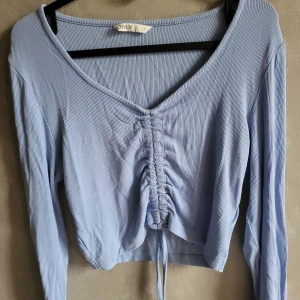 Ljusblå ribbad topp från ONLY - Säljer en ljusblå ribbad topp från ONLY med v-ringning och knytning framtill. Toppen har långa ärmar och en croppad passform, perfekt för en avslappnad stil.