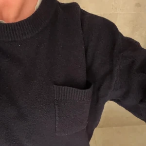 Mörkblå stickad tröja med ficka - Säljer en mörkblå stickad tröja med en praktisk ficka på bröstet. Tröjan har en ribbad krage och långa ärmar, perfekt för kyligare dagar. Den har en avslappnad passform som ger en bekväm känsla.