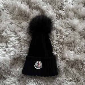 Svart stickad mössa med pälsboll - Snygg svart stickad mössa från Moncler med en stor pälsboll på toppen. Mössan har en ribbad design och ett märke framtill. Perfekt för kalla vinterdagar.