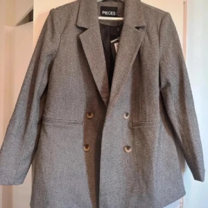 Grå kavaj från Pieces - Snygg grå kavaj från Pieces med dubbelknäppning och klassisk krage. Perfekt för en stilren look. Passar bra till både vardag och mer formella tillfällen.