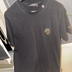 Svart t-shirt från Philipp Plein - Snygg svart t-shirt från Philipp Plein med ett coolt emblem på bröstet. Den har korta ärmar och en klassisk rund halsringning. Perfekt för en stilren look.