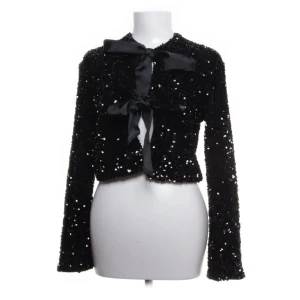 Svart paljettjacka med rosett - Snygg svart paljettjacka med långa ärmar och en elegant rosett framtill. Perfekt för att glittra på festen. Jackan är croppad och har en stilren design.