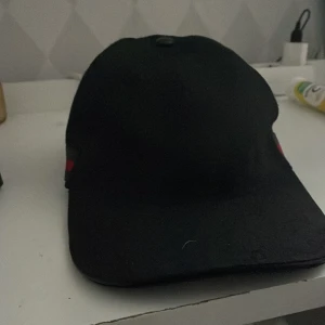 Gucci keps svart - Snygg svart keps från Gucci med gröna och röda ränder på sidan. Kepsen har en justerbar rem baktill och ett diskret mönster med märkets logotyp. Perfekt för en stilren look.