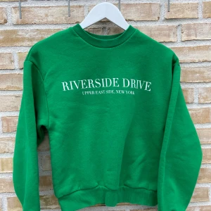 Grön sweatshirt från Gina Tricot🌟 - Säljer en grön sweatshirt från Gina Tricot med texten 'Riverside Drive Upper East Side, New York' tryckt på framsidan. Tröjan har en klassisk rund hals och ribbade muddar vid ärmslut och nederkant. Perfekt för en avslappnad men snygg stil.