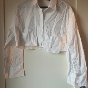 Vit croppad skjorta från Zara - Säljer en stilren vit croppad skjorta från Zara. Skjortan har långa ärmar och knappar framtill. Perfekt för en trendig look.