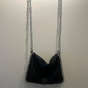 Svart axelväska från Tiamo - Snygg svart axelväska från Tiamo med kedjedetaljer och dragkedja. Perfekt för att ge din outfit en edgy touch. Väskan har en praktisk storlek och är lätt att bära med sig.