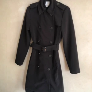 Svart trenchcoat från H&M - Snygg svart trenchcoat från H&M med klassisk dubbelknäppning och bälte i midjan. Perfekt för höst och vår. Jackan har långa ärmar och en stilren krage.