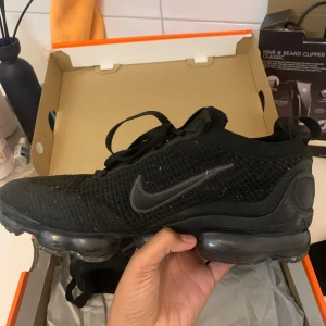 Svarta Nike Air Vapormax 2021 - Säljer ett par svarta Nike Air Vapormax 2021 med en stilren design och bekväm passform. Skorna har en stickad ovandel och synlig Air-dämpning i sulan för extra komfort. Perfekta för både träning och vardag. Använde dem 2 gånger men passade inte direkt. Visar inget tecken på användning 