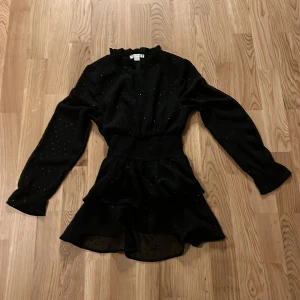 Svart glittrig klänning från Gina Tricot - Snygg svart klänning från Gina Tricot med glittriga detaljer. Klänningen har långa ärmar och en volangdetalj i midjan som ger en fin siluett. Perfekt för festliga tillfällen!