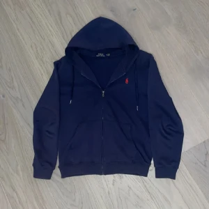 Mörkblå hoodie från Polo Ralph Lauren - Säljer en stilren mörkblå hoodie från Polo Ralph Lauren med dragkedja och röd broderad logga på bröstet. Perfekt för en avslappnad look med justerbar huva och långa ärmar. Storleken är M men passar S-XS 