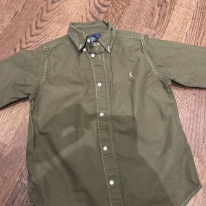 Olivgrön skjorta från Ralph Lauren st 7 år  - Säljer en olivgrön skjorta från Ralph Lauren med klassisk krage och knappar framtill. Skjortan har långa ärmar och en liten broderad logotyp på bröstet. Perfekt för en stilren look. Oanvänd men ändå nga fläckar. Skickar bild. 