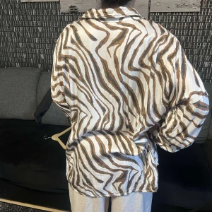 Brun- och vitmönstrad skjorta - Snygg och luftig skjorta med brunt och vitt zebramönster. Skjortan har långa ärmar och knappar framtill, perfekt för en avslappnad stil.