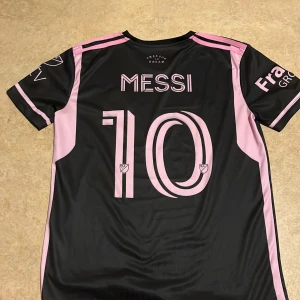 Messi Inter Miami tröja - Snygg svart fotbollströja med rosa detaljer från Adidas. Tröjan har korta ärmar och ett tryck med nummer 10 och namn på baksidan. Perfekt för fotbollsfans!