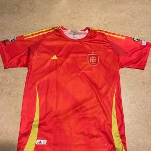 Röd Spanien tröja - Säljer en röd kortärmad fotbollströja från Adidas med gula detaljer. Tröjan har Spaniens landslagsemblem på bröstet och är perfekt för fotbollsfans. Den har även sponsorloggor på ärmarna.