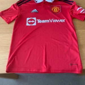 Röd Manchester United fotbollströja från Adidas - Säljer en röd Manchester United fotbollströja från Adidas med korta ärmar och vit krage. Tröjan har klubbens emblem på bröstet och sponsorn TeamViewer tryckt på framsidan. Perfekt för fans av laget!