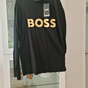 Svart långärmad tröja från Boss - Säljer en stilren svart långärmad tröja från Boss med stort tryck på bröstet. Perfekt för en avslappnad men ändå snygg look. Tröjan är gjord i mjukt material och har en klassisk passform.